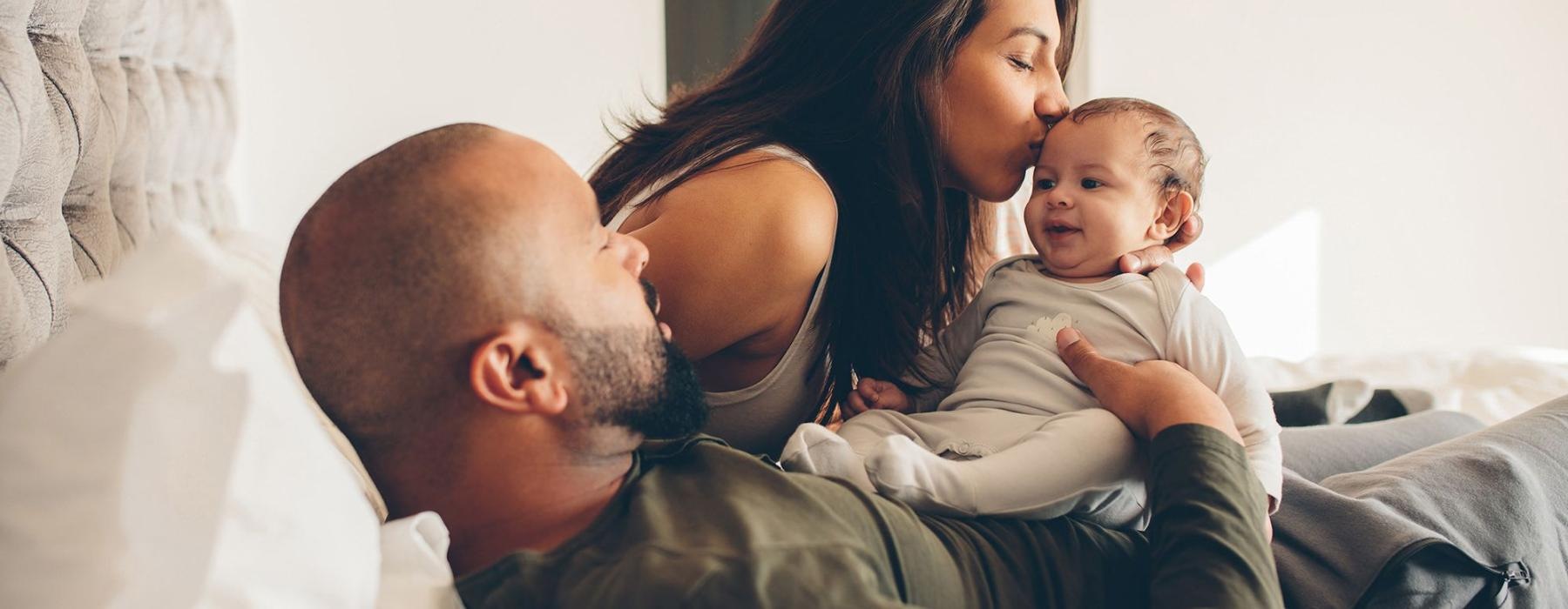 mother kisses her infant while father lies in bed and holds him